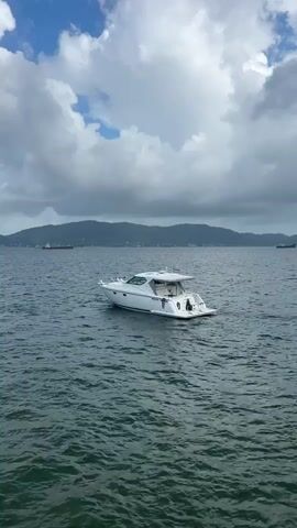 A white motor yacht is anchored in the ocean. The yacht is facing to the right of the frame. There is a body of water in the foreground. In the background, there are mountains and a large cargo ship. The sky is cloudy.