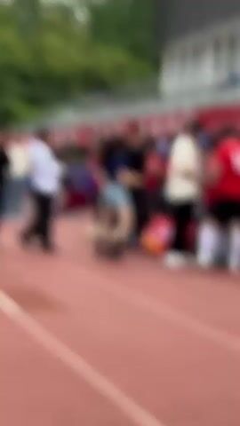 The video shows a group of young men in red soccer uniforms standing on a red running track, apparently receiving an award or congratulations from two men. They are standing in a huddle, shaking hands with the men. One man is dressed in a black Chinese-style outfit, the other is in a white button-down shirt and black pants. There is a small table with a trophy and what appears to be a banner behind the men. In the background, there are people standing along a railing in front of a building and bleachers. The event appears to be taking place on a college campus, with signs visible in Chinese. Trees can be seen in the background.