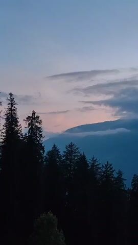A time-lapse video shows a dark silhouette of a forest in the foreground with mountains in the background as the sky transitions from twilight to a darker shade. Clouds drift across the sky, and a faint glow from the sun can be seen peeking through the clouds behind the mountains. The video pans slightly upwards to reveal more of the sky.