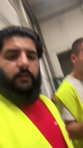 Two men are in a warehouse. The man on the left is wearing a red shirt and a yellow safety vest. He has dark hair and a beard. The man on the right is wearing a grey t-shirt and a yellow safety vest. He has short hair and is smiling. He is holding a phone in his right hand and making a gesture with his left hand. In the background, there is a large green dumpster. The lighting is dim.
