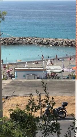 The video pans across a view of a beach and the ocean on a sunny day. In the foreground, there is a blurred green plant in front of a motorbike parked on the side of a road. Beyond the road is a beach with several people sunbathing and walking. There are a few small structures on the beach, possibly changing rooms or cafes, and umbrellas providing shade. A breakwater made of large rocks separates the beach from the deeper ocean, which is a vibrant turquoise color. The sky is clear and blue.