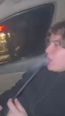 A young, white male in a black hoodie is seen from the side inside a car. He is holding a long, black object and exhaling a large puff of white smoke from his mouth. The window behind him is wet and shows a blurred view of a building exterior with bright lights and a red sign. He takes another inhale from the black object and then exhales more white smoke. The car appears to be in motion, and the light outside changes, indicating movement.