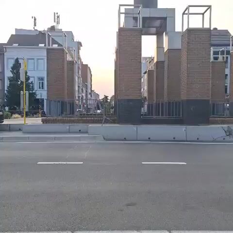 The video displays a street scene with a modern sculpture or monument in the center, made of brick columns and metal frames, with a tall, dark structure on top. The sculpture is situated in a public space, possibly a square or intersection, with a road in the foreground and buildings lining the street in the background. The sky is visible with some clouds and hints of sunset colors. There are no people or vehicles clearly visible in the frame. The lighting suggests it is either early morning or late afternoon. The overall impression is an urban setting with a prominent artistic structure.