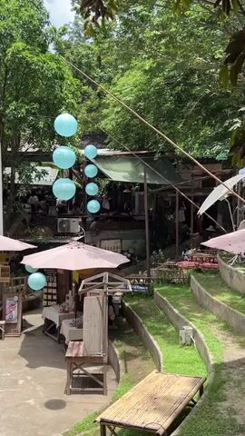 The video shows an outdoor seating area at a cafe or restaurant. There are many trees and lush greenery surrounding the space. Hanging blue paper lanterns are strung across the area. In the foreground, there is a bamboo seating bench on a tiered grassy slope. Further back, there are tables with umbrellas, food stalls, and people sitting and dining.