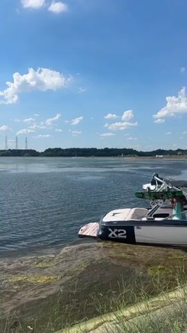 The video shows a wide expanse of calm water under a partly cloudy blue sky. In the foreground, a wakeboard boat with the "X2" logo is partially submerged at the edge of the water, resting on a ramp or shallow area. The water near the shore is murky and filled with green algae and debris, including a partially submerged log. In the distance, the opposite shore is lined with trees. Power lines and distant structures can be seen on the horizon. There are no people visible in the video. The overall scene depicts a serene, but slightly unkempt, waterside location on a sunny day.