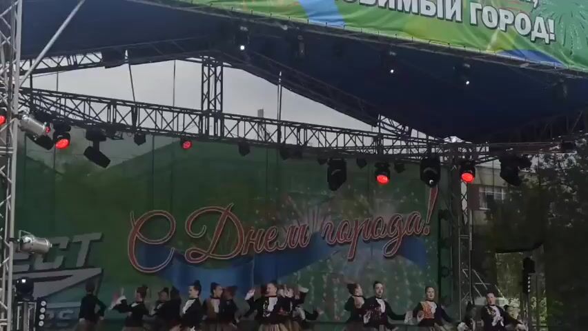 A group of young girls is performing a dance routine on an outdoor stage. They are dressed in black tops and fluffy, layered skirts. The stage is set up with a large banner in the background that reads "С Днем города!" which translates to "Happy City Day!". The banner also features a graphic of fireworks. Several spotlights and stage lights are visible above the banner. In the foreground, the heads of an audience are visible, suggesting the performance is taking place at a public event. The overall atmosphere appears festive. The lighting suggests it is daytime, possibly overcast, given the diffused light. The presence of a stage and banner indicates a celebration or festival in a town or city.