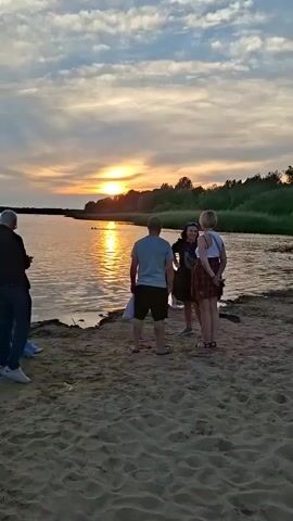 The video opens on a sandy beach at sunset. A group of people are gathered near the water's edge. In the background, trees line the horizon. The video pans across the beach, showing more people, some sitting and some standing. A kite is visible in the sky. The scene shifts to show two people standing on the sand, facing away from the camera. The video concludes with a wide shot of the beach, with many people scattered across the sand, some appearing to be around a bonfire.
