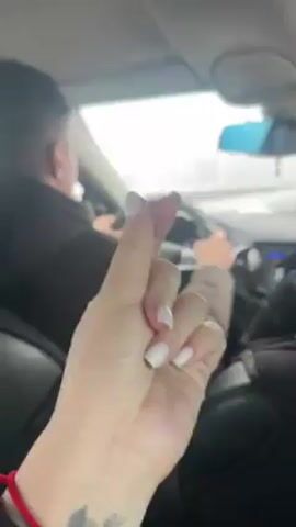 The video shows the inside of a car during the daytime. A person in the passenger seat holds up their hand to make a small heart symbol. This hand has white nail polish, a red string bracelet, and a tattoo on the wrist. A driver is visible in the background, holding the steering wheel and looking forward. The weather appears overcast or snowy outside the car windows.
