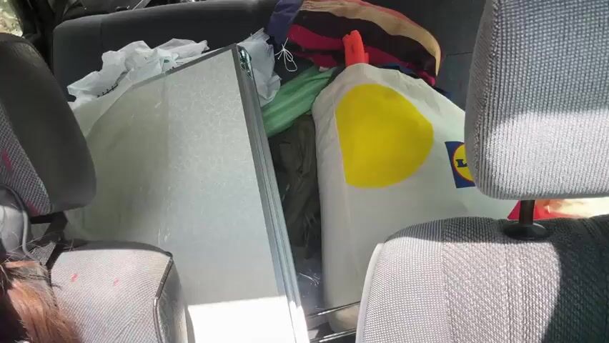 The video begins inside a car, showing the back seats filled with various items. A person's dark brown hair is visible on the left side of the frame. The car is packed with what appears to be camping gear or personal belongings, including a folded mattress or similar flat object, a large colorful striped bag, a green sleeping bag or folded fabric, and a white pillow with a yellow circular logo that resembles the Lidl brand. The interior of the car is dimly lit compared to the exterior.

The camera then tilts upwards, revealing the view through the car's windows. The scene shifts to a bright, sunny sky with scattered white clouds. Lush green trees frame the sky, with their branches and leaves visible against the blue. The sun is shining intensely, creating lens flare and a bright glow in the sky. The overall impression is a view from a moving vehicle on a clear, sunny day, looking out towards the sky and surrounding foliage.