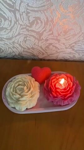 A hand holds a lit match near a white, peony-shaped candle that is already lit. The match is positioned to the left of the candle's flame. To the right of the white candle is a pink, peony-shaped candle, also lit, with a small red heart-shaped object behind it. The items are arranged on a pink and white marbled tray. The tray sits on a wooden surface. Behind the tray is a wall with patterned wallpaper. The lighting in the scene is dim, with the flames of the candles providing illumination.