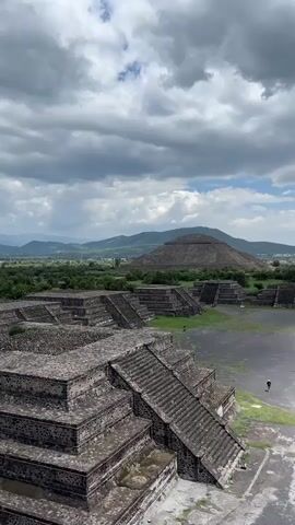 This media file showcases an aerial view of the Teotihuacan archaeological site in San Francisco Mazapa, Mexico. The scene is dominated by ancient stone structures, including the imposing Pyramid of the Sun in the background, and various plazas and smaller pyramids in the foreground. The architecture is characterized by its stepped design and rough stone construction, indicative of Mesoamerican civilizations.

A few people are visible walking around the site, dwarfed by the scale of the ruins, suggesting it is open to tourists. The setting is vast and open, with lush green vegetation surrounding the ancient city, and rolling hills and mountains in the distant background.

The weather appears to be overcast with dramatic, heavy clouds interspersed with patches of blue sky, hinting at a possibly humid or recently rainy day. The time of day is difficult to ascertain precisely due to the cloud cover, but it appears to be daytime, likely late morning or afternoon.

The overall emotion evoked is one of awe and historical significance, highlighting the grandeur of the ancient civilization that built these structures. There is no visible text in the media file.