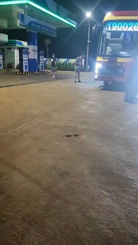 The video shows a nighttime scene at a gas station in Xuân Long, Vietnam. Two people are standing on the concrete lot, and a large bus is parked under a green and white gas station canopy. The bus has the number "19002679" displayed prominently on its front. Another vehicle with bright headlights is approaching the gas station from the right, its lights illuminating the wet pavement. The overall lighting suggests it is dark outside, and the wet ground could indicate recent rain. The gas station itself has illuminated signage and pillars, with additional signs visible in the background. The atmosphere appears casual, with people engaged in typical gas station activities. The visible text on the bus and the gas station signage provides specific identifying information.
