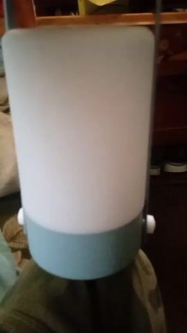 A light fixture is held up by a person. The light fixture is cylindrical with a translucent white shade and a light blue base. A handle extends from the top of the fixture. The background is blurred and appears to be an indoor setting with furniture and a dark floor. The light fixture is turned on at the end of the video, illuminating brightly.