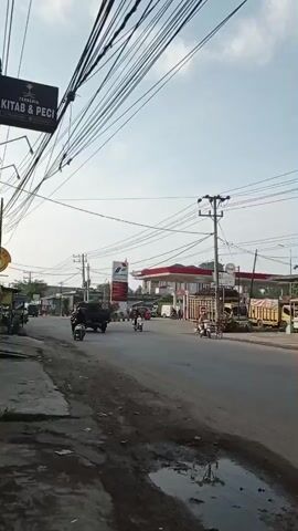 The video shows a street scene in what appears to be a commercial area, possibly in Southeast Asia, given the signs and architecture. The camera pans from left to right, capturing various buildings and vehicles.

At the beginning, a gas station with the Pertamina logo is visible. Several motorcycles and a truck are present. A sign that says "Baju & PECI" is also visible. The road has puddles of water, indicating recent rain or a drainage issue. Overhead power lines are a prominent feature throughout the video.

As the camera continues to pan, more shops and buildings come into view. Some of these appear to be clothing stores or general merchandise shops, with signs in a foreign language. Several motorcycles are seen moving along the road, and some are parked. A white van is also visible.

Further along the street, buildings with multiple stories are shown, some with arched windows and decorative elements. More shops are lined up, with some of their entrances covered by roller shutters. One shop has a sign that reads "NAHYA MOTOR" and has several motorcycles covered with tarps parked in front.

The sky is partly cloudy, and the lighting suggests it is daytime, likely late morning or early afternoon. The overall impression is of a busy, developing urban or semi-urban environment.
