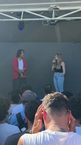 This media file captures a dynamic moment on a stage, likely during an outdoor event. Two individuals are the primary subjects: on the left, a man with dark hair, wearing a red cardigan over a white t-shirt and dark pants, stands holding a microphone. To his right, a woman with blonde hair, dressed in a black tank top and light blue jeans, also holds a microphone and gestures with her free hand, engaging with the audience.

They are positioned on a raised stage with a dark backdrop and a white overhead structure with lighting fixtures. Below the stage, a crowd of people is visible, their backs turned to the camera, indicating they are facing the speakers. The focus is on the interaction between the two individuals on stage, suggesting a Q&A session, an interview, or a presentation.

The lighting suggests it's daytime, possibly late afternoon, given the bright ambient light and the shadows. The presence of a crowd and the stage setup point towards a public gathering or festival. The individuals appear engaged and possibly enthusiastic, judging by their postures and the woman's gesture.

Although no text is explicitly legible on clothing or the backdrop, the event could be related to a film or entertainment event, given the celebrity-like appearance of the individuals and the enthusiastic audience. The context provided mentions Cesena, Italy, which helps localize the event to that specific city. The overall scene conveys a sense of community and shared experience.