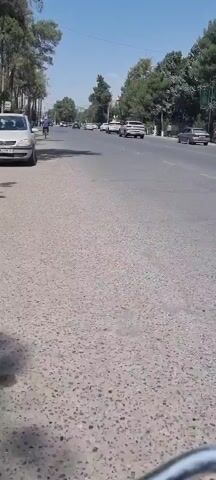 A street view video taken from a sidewalk. The camera is moving forward at a walking pace. The street is paved and appears to be a main road with multiple lanes of traffic. Cars are visible driving in the distance. On the left side of the road, a parked white car is visible. Trees line the sidewalk and the opposite side of the road, casting shadows on the pavement. The sky is clear and blue.