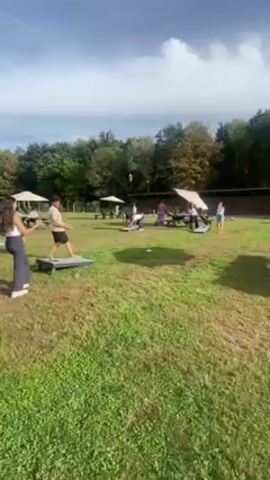 The video shows a grassy area with a group of people playing cornhole. Two cornhole boards are visible in the foreground, with people standing by them. In the background, there are more people playing at several other cornhole boards, along with picnic tables under umbrellas. The sky is partly cloudy and there are trees in the background. A woman is throwing a cornhole bag at the board.