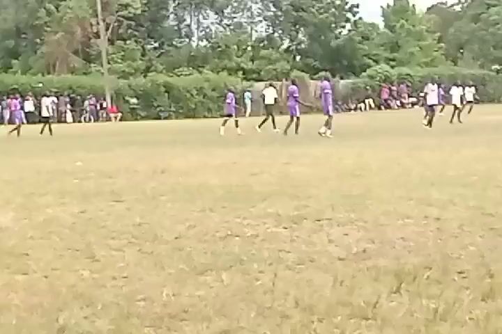 The video shows a soccer game in progress on a grassy field. Players in purple uniforms and players in white uniforms are running, kicking, and chasing a soccer ball. There is a soccer goal visible on one end of the field. A building with multiple windows is visible behind the field. People are sitting on the sidelines watching the game. The sky is overcast.