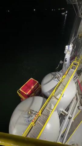 The video shows an industrial offshore platform at night, likely near Marina di Ravenna, Italy. The camera pans upwards, revealing a complex structure of metal frameworks, walkways, and stairs, all brightly illuminated by numerous lights. Several large, grey, cylindrical tanks are visible on one of the lower levels of the platform. Adjacent to the platform, on the dark water, a red container or crate is seen. The water reflects the platform's lights, and distant lights are visible on the horizon, possibly from other vessels or the shore. The scene is dominated by the intricate industrial architecture against the dark night sky. No people or visible text are discernible. The video captures the scale and illumination of the offshore facility.
