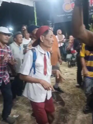 The video depicts an outdoor event, possibly a performance or celebration, under a tent. In the foreground, a person in a black shirt is seen with their arm raised, facing away from the camera. To the left, a person in a white school uniform with a red tie and a red cap is looking towards the center. Behind them, other individuals are visible, including a person in an orange shirt and another in a yellow and blue striped shirt.

The background features a stage with musicians, most of whom are wearing yellow shirts. Musical instruments, including guitars and keyboards, are present on the stage. A red carpet covers the stage area. Several water bottles and what appear to be empty cups are scattered on the red carpet in front of the stage. The overall environment suggests a lively gathering with a focus on entertainment.