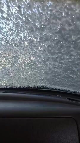 The video provides a POV shot from inside a car, looking through the windshield. The windshield is covered in soap bubbles and water, suggesting the car is being washed. The dashboard and steering wheel are visible, indicating the driver's perspective. A rearview mirror hangs from the windshield, and a decorative item with red and blue stripes is attached to it. The overall scene is of a car going through a car wash.