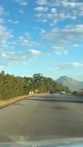 A car drives down a paved highway with a mountain in the distance beneath a blue sky with puffy white clouds. The roadside is lined with trees and bushes. Other cars travel on the highway, moving in the same direction.