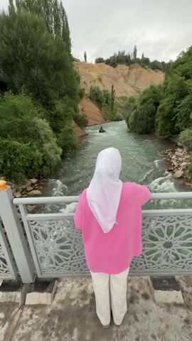 The video shows a person wearing a white hijab and a pink sweater standing on a bridge overlooking a flowing river. The river is surrounded by lush green trees and a hilly landscape with brown soil. There is a person in a black raft or kayak in the river. The sky is cloudy. The person on the bridge is facing away from the camera, looking at the river. The bridge has a white railing with an intricate pattern.