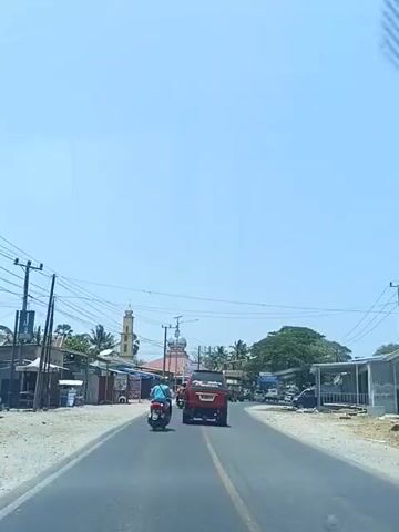 A motorcycle with a rider wearing a blue shirt and a backpack drives down a paved road in a rural area. The motorcycle is followed by a red SUV. On the left side of the road, there are small shops and a mosque with a tall minaret. On the right side of the road, there are more buildings and trees. The sky is clear and blue, and the sun is shining brightly. The video is filmed from the perspective of someone in another vehicle.