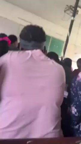 The video shows a crowded classroom from the perspective of someone in the back. People are mostly standing, and they seem to be facing the front where a young woman is standing in front of a green chalkboard. Most of the people in the room have dark hair and dark skin. The room appears old and is sparsely furnished. A black and white pole runs from the floor to the ceiling. There is writing on the chalkboard behind the young woman. Most of the students have a notebook or paper in their hands. There is a loud noise in the background.