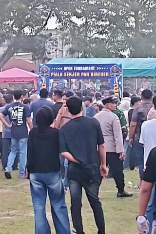 The video shows an outdoor event with a large crowd of people gathered around a stage or booth that says "OPEN TOURNAMENT PIALA SENJEN PAN BIREUEN". There are tents set up, one pink and one blue, and trees in the background. Many people are standing and looking towards the stage. Some individuals are wearing uniforms, including military camouflage and police. The ground is covered in grass. The overall atmosphere appears to be festive and crowded.