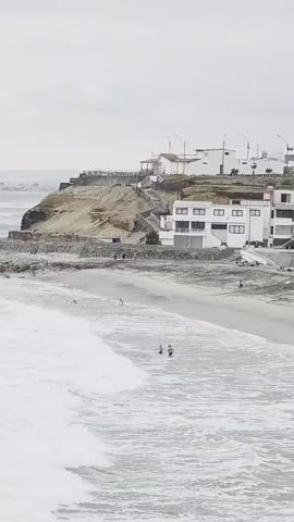 The video shows a view of a beach with white buildings on a cliff overlooking the ocean. There are waves crashing on the shore, and a few people are visible in the water and on the sand. The sky is overcast.