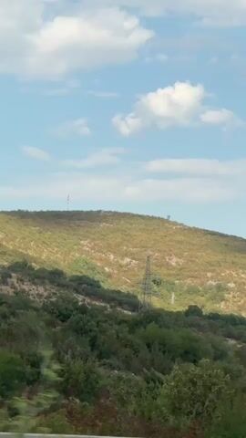 The video pans across a dry, hilly landscape under a partly cloudy sky. Sparse vegetation covers the hillsides, with patches of green and brown. In the distance, there are several tall, metal towers that appear to be electricity pylons. A few small buildings or structures are visible on the crest of one of the hills. In the foreground, the tops of green trees and bushes are visible, and a traffic sign is partially seen on the left. The overall impression is a rural, possibly arid environment.