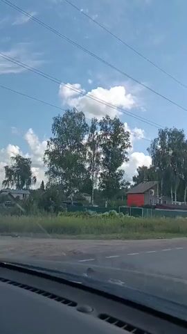 The video shows a view from inside a vehicle, looking out at a rural landscape on a sunny day with scattered clouds. The vehicle is driving on a paved road, indicated by lane markings. On the left side of the road, there are trees, grass, and a few houses. On the right side, there are also trees and a field of grass. Power lines are visible stretching across the sky. A dark-colored car is driving ahead of the vehicle, and another dark-colored vehicle is visible further down the road. The scene suggests travel through a rural or suburban area.