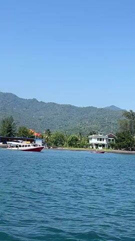 The video shows a red and white boat moving from right to left across a body of water. In the background is a green, forested hillside. On the right side of the screen is a white, multi-story building. On the left side of the screen, there is a sheltered area with people walking and sitting underneath it. The sky is clear blue.
