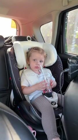 A little girl, around 2 years old, is sitting in a black and white Joie car seat in a car. She is wearing a white T-shirt, beige leggings, and a white bib with cloud shapes on it. Her hair is pulled back in a small ponytail. She is holding a clear plastic cup with a straw and drinking from it. The drink appears to be a pink liquid. The car's interior is visible, including the black leather seats and a window with sunlight shining through. Outside the window, there is a house.