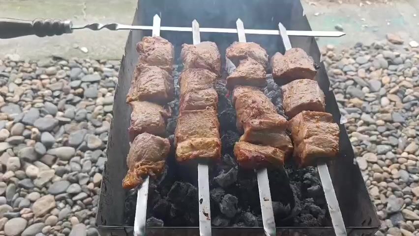 The video shows four skewers of meat cooking over charcoal in a grill. The meat appears to be cut into cubes and is browning on all sides. Smoke rises from the grill, indicating that the meat is actively cooking. The grill is placed on a surface covered with small, grey pebbles. In the background, a portion of a light-colored wall or surface is visible. The lighting suggests an outdoor setting, likely during the daytime. There are no people visible in the frame. The focus is entirely on the grilling meat.