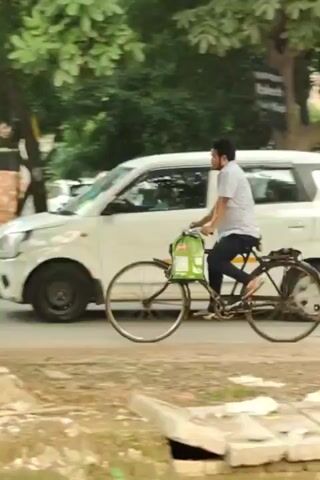 A man is riding a bicycle on a road. He is wearing a white shirt and dark pants. The bicycle has a green bag attached to the front. There is a white car on the left side of the road and trees on the right. The ground is covered with dirt and leaves. The weather appears to be daytime and sunny. The man is pedaling the bicycle and moving forward.