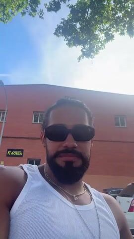 A man with a beard, long dark hair tied back, sunglasses, and a white tank top records himself from the chest up, looking directly at the camera. He wears multiple silver necklaces. The camera pans right, revealing two other men sitting on a bench beside him; one in a patterned shirt is looking at a white phone, and another in a white t-shirt is also engaged with a device. A fourth man in a white t-shirt and light pants walks into the frame and stands in the background. The scene is outdoors with a clear sky, trees, a dirt ground, a wooden fence, and brick buildings in the background, one partially labeled "ACISA". Several cars are parked behind the individuals.