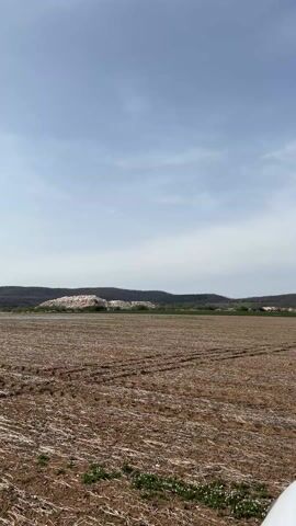 The video shows a view from inside a white vehicle driving through a plowed field on a partly cloudy day. The field is brown and appears recently tilled, with visible tire tracks. In the distance, there are rolling hills and trees. The windshield wipers of the vehicle are visible. The sky is light blue with patches of white clouds. There are no people visible in the video.