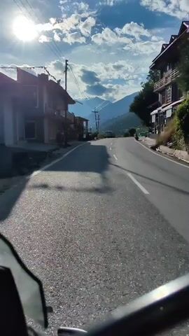 This is a first-person perspective video of a motorcycle ride on a paved mountain road. The video captures a scenic view of a valley with mountains in the background and the sun shining brightly in a partly cloudy sky. The road curves to the right, and on the left side of the road, there are buildings and some vegetation. On the right side, the mountain slope is covered with trees and vegetation. Several vehicles, including cars and possibly other motorcycles, are visible on the road and parked along the side. The video is shaky, suggesting movement and an active journey.