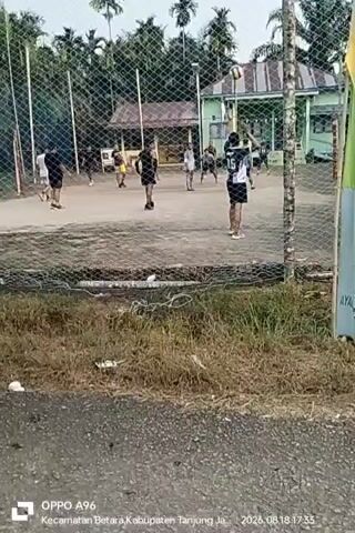 The video shows a group of young people playing volleyball on an outdoor court. The court appears to be made of dirt or sand, and it is surrounded by a chain-link fence. In the background, there are trees and a building that looks like a school or community center. The players are dressed in casual athletic wear. The action captured in the video is a volleyball game in progress, with players moving around the court, hitting the ball, and attempting to score. The lighting suggests it is either early morning or late afternoon. The timestamp on the video indicates it was recorded on August 18, 2026, at 17:33 in Betara Subdistrict, Tanjung Jabung Regency.