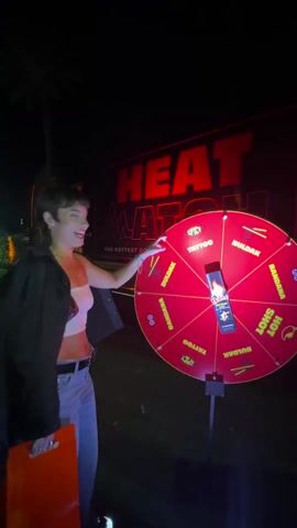 A woman stands in front of a red wheel with yellow lines, a game labeled "Heat Watch, the Hottest". She is wearing a white top, blue jeans, and a black jacket, and she has short brown hair. She spun the wheel, which has spaces labeled "Spicy", "Tattoo", "Bandana", "Buldak", and "Hot Shot". The wheel is mounted on a stand and is at nighttime. In the background, there is a large sign that reads "Heat Watch". The wheel lands on "Tattoo".