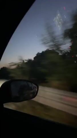 A video captures the view from inside a moving vehicle at dusk. The camera faces the side window, showing the passing scenery. A side mirror is visible in the foreground, reflecting the interior of the car and the road behind it. The sky transitions from a deep blue at the top to a lighter shade near the horizon, with a faint hint of orange or pink indicating the setting sun. Silhouetted trees and bushes line the sides of the road, blurred due to the vehicle's motion. Streaks of light from passing headlights or streetlights are visible in the distance, further emphasizing the movement. A small digital display or object is reflected on the windshield at the top right of the frame. The overall atmosphere is one of travel and the quiet beauty of twilight.
