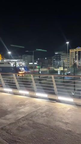 At night, the video shows a person walking across a bridge illuminated by several street lights. A large ferry is docked in the background. The person then enters a glass-walled structure and rides down an escalator. As the escalator descends, a woman wearing a white jacket, denim shorts, a black backpack, and sneakers walks onto the escalator. The video ends with the escalator descending toward street level.
