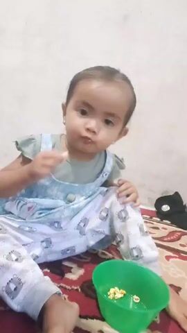 A young child is sitting on a patterned rug. The child is wearing a light blue overall with white pants patterned with cartoon lions. The child has dark hair and is looking at the camera. The child is eating something, and a green bowl containing food is in front of the child. The background is a plain wall.