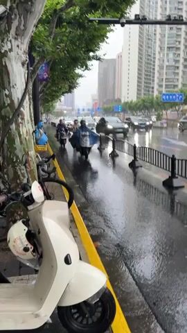 The video shows a street scene during a rain shower. In the foreground, a white electric scooter is parked on the sidewalk next to a tree. The sidewalk is lined with a yellow curb. Further down the street, several people are riding scooters and motorcycles, most of them wearing raincoats. Cars are driving on the wet road, creating splashes. Traffic lights and street signs are visible in the background, along with tall buildings. The overall atmosphere is overcast and wet due to the rain.