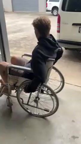 A young man with curly, light-brown hair sits in a wheelchair. He wears a black hoodie with a white logo on the chest, khaki shorts, and brown work boots. He is indoors on a concrete floor, with a closed garage door behind him. He turns to face the camera and says, "Hey Josh. Hey Josh. Jet lags." He then turns and looks to the side. A white van is partially visible in the background. Another person in work boots is briefly visible in the background as well.