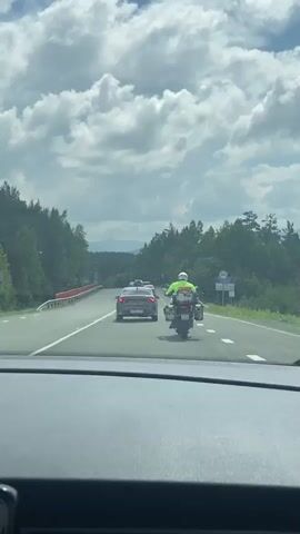 The video captures a view from inside a car driving on a highway. The car is following a motorcycle with a rider wearing a bright yellow jacket and a helmet. The motorcycle is in the right lane, and behind it, several cars are also traveling in the same direction. The road is flanked by lush green trees, and the sky is filled with dramatic white clouds against a blue backdrop. In the distance, hills or mountains can be seen. A red barrier is visible along the side of the road, and a blue road sign is posted on the right. The perspective suggests the camera is mounted on the dashboard or windshield of the car, looking forward through the front window.