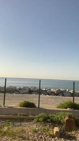 The video shows a view of the ocean through a chain-link fence. There are rocks piled along the shoreline, and the water appears calm under a clear sky. The camera is moving, suggesting it's being filmed from a vehicle. The foreground is gravelly with some sparse vegetation. There are no people visible.