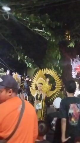 A nighttime outdoor event is captured in this video. In the foreground, a person wearing an orange shirt and a baseball cap is partially visible, looking to the left. Behind them, a crowd is gathered. To the left of the frame, a person in an elaborate golden costume with a circular headdress is visible, and to the right, a large, illuminated dragon decoration is present. In the upper part of the frame, a person in a lighter colored costume, possibly a deity or performer, is suspended by wires among tree branches. The environment appears to be a street with trees and overhead wires.